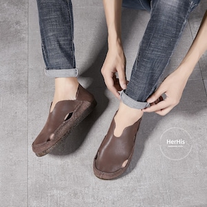 May include: Brown leather slip-on shoes with a cut-out design on the top. The shoes have a brown leather sole and are worn by a person wearing jeans.