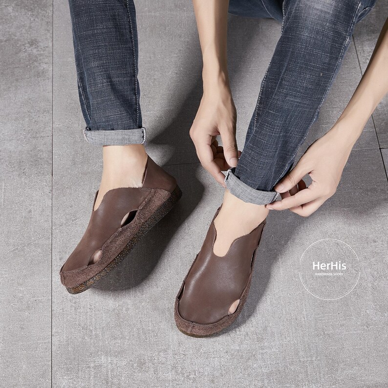 May include: Brown leather slip-on shoes with a cut-out design on the top. The shoes have a brown leather sole and are worn by a person wearing jeans.