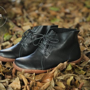 May include: A pair of black leather ankle boots with laces. The boots have a brown sole and are sitting on a bed of fallen leaves.