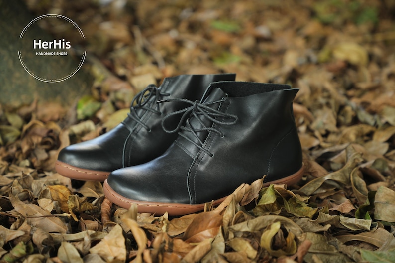 May include: A pair of black leather ankle boots with laces. The boots have a brown sole and are sitting on a bed of fallen leaves.