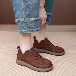 May include: Brown leather shoes with a rounded toe and a thick, light brown sole. The shoes have laces and a small tab at the heel. The person is wearing blue jeans.