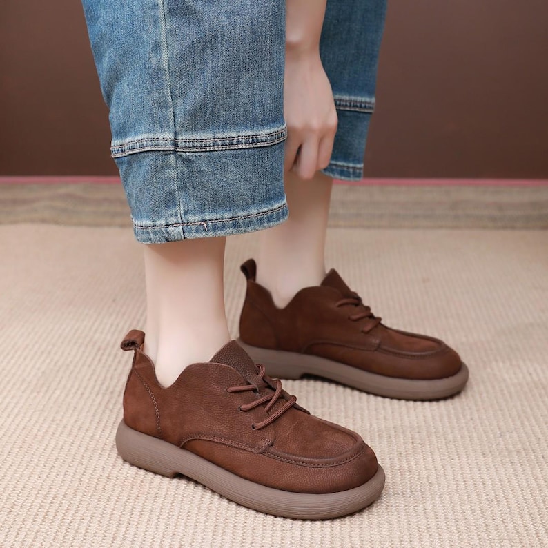 May include: Brown leather shoes with a rounded toe and a thick, light brown sole. The shoes have laces and a small tab at the heel. The person is wearing blue jeans.