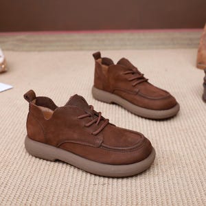 May include: A pair of brown leather shoes with a rounded toe and a thick, light brown sole. The shoes have laces and a low-cut design, with a small pull tab at the heel. The shoes are made of a textured leather material.