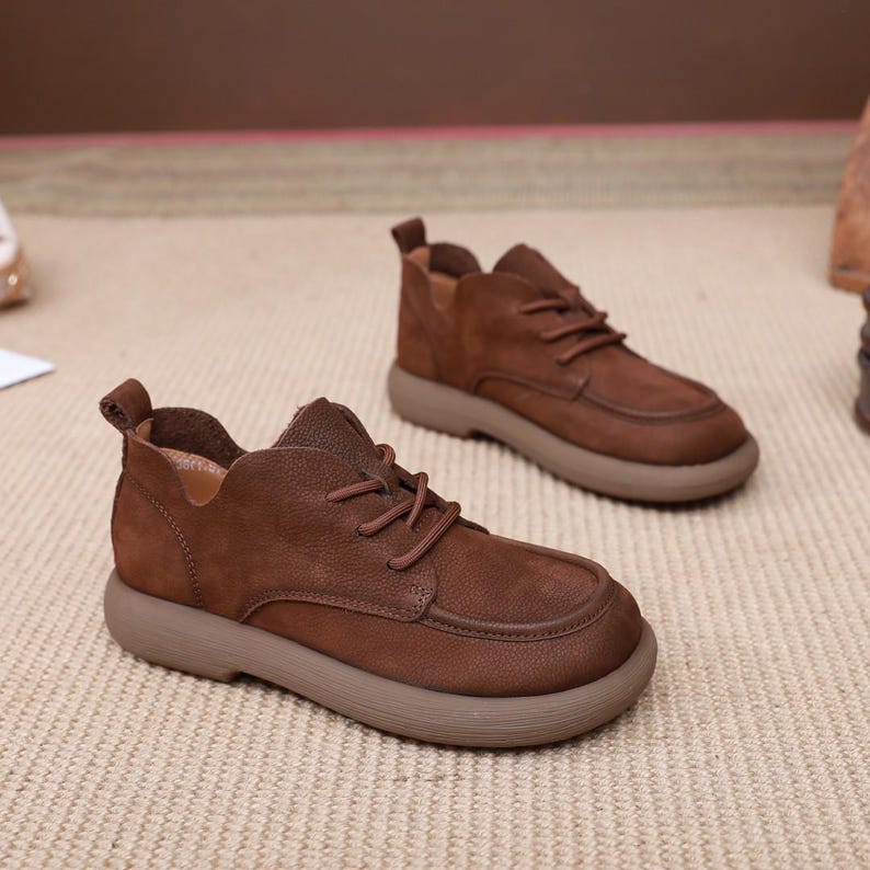 May include: A pair of brown leather shoes with a rounded toe and a thick, light brown sole. The shoes have laces and a low-cut design, with a small pull tab at the heel. The shoes are made of a textured leather material.