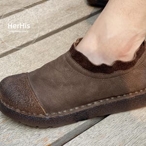 Op de afbeelding: Bruine leren instapper met een ronde neus en een textuurzool. De schoen heeft een donkerbruine rand rond de opening en de teen. Handgemaakt, met de tekst "HerHis HANDMADE SHOES" in een cirkelvormig logo.