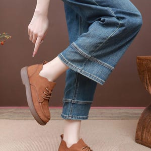 May include: Brown leather shoes with laces and a thick sole. The shoes are worn with blue denim jeans. The image shows a person pointing at the shoes. The shoes appear to be a casual style.