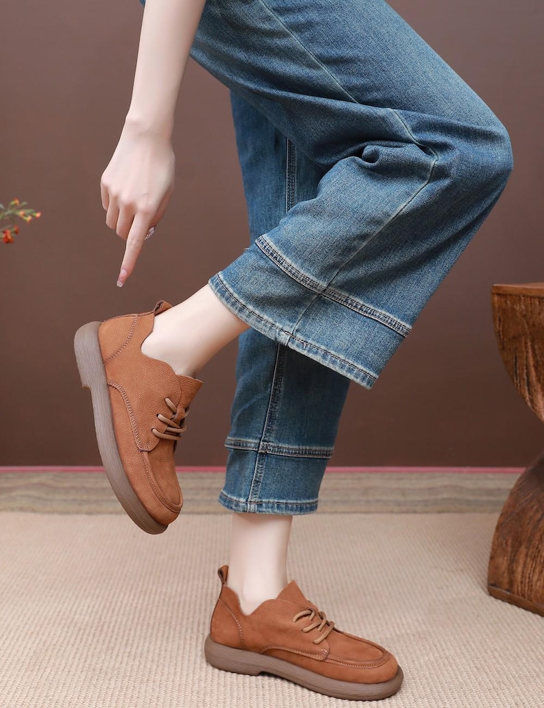 May include: Brown leather shoes with laces and a thick sole. The shoes are worn with blue denim jeans. The image shows a person pointing at the shoes. The shoes appear to be a casual style.
