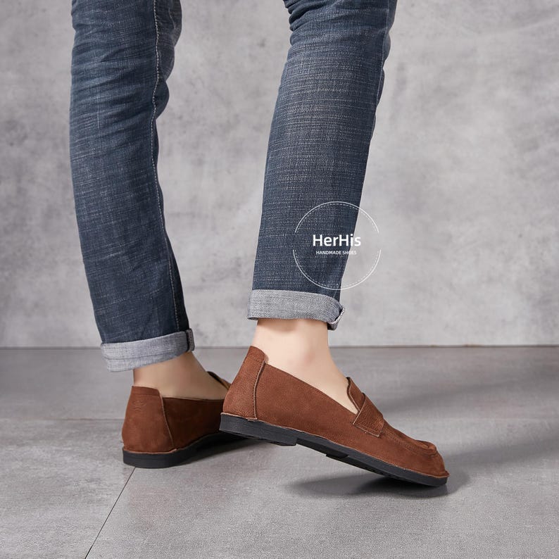 May include: Brown leather loafers with a slip-on design. The shoes have a slightly pointed toe and a low heel. The shoes are worn by a person wearing blue jeans.
