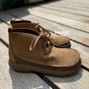 May include: A pair of brown leather ankle boots with laces. The boots have a suede-like texture and a thick sole. The brand name "HerHis Handmade Shoes" is visible on the label.