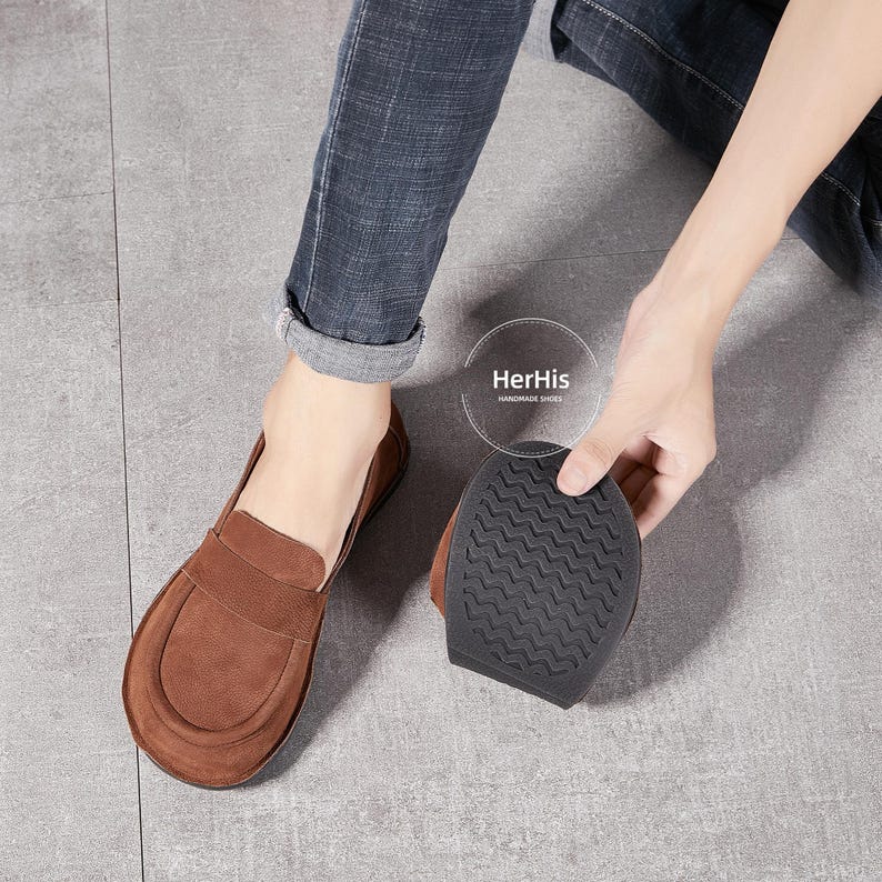 May include: Brown leather loafers with a suede finish and a black rubber sole. The sole has a textured pattern. The shoes are handmade by HerHis.