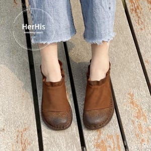 Op de afbeelding: Bruine leren schoenen met een ronde neus en een gerimpelde kraag. De handgemaakte schoenen hebben een distressed afwerking. De afbeelding toont ook de tekst "HerHis HANDMADE SHOES".