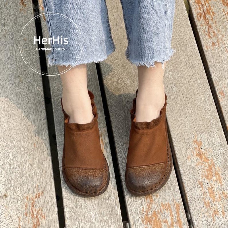 Op de afbeelding: Bruine leren schoenen met een ronde neus en een gerimpelde kraag. De handgemaakte schoenen hebben een distressed afwerking. De afbeelding toont ook de tekst "HerHis HANDMADE SHOES".