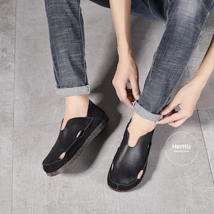 May include: A pair of black leather slip-on shoes with a cut-out design. The shoes have a rubber sole and are worn by a person wearing jeans.