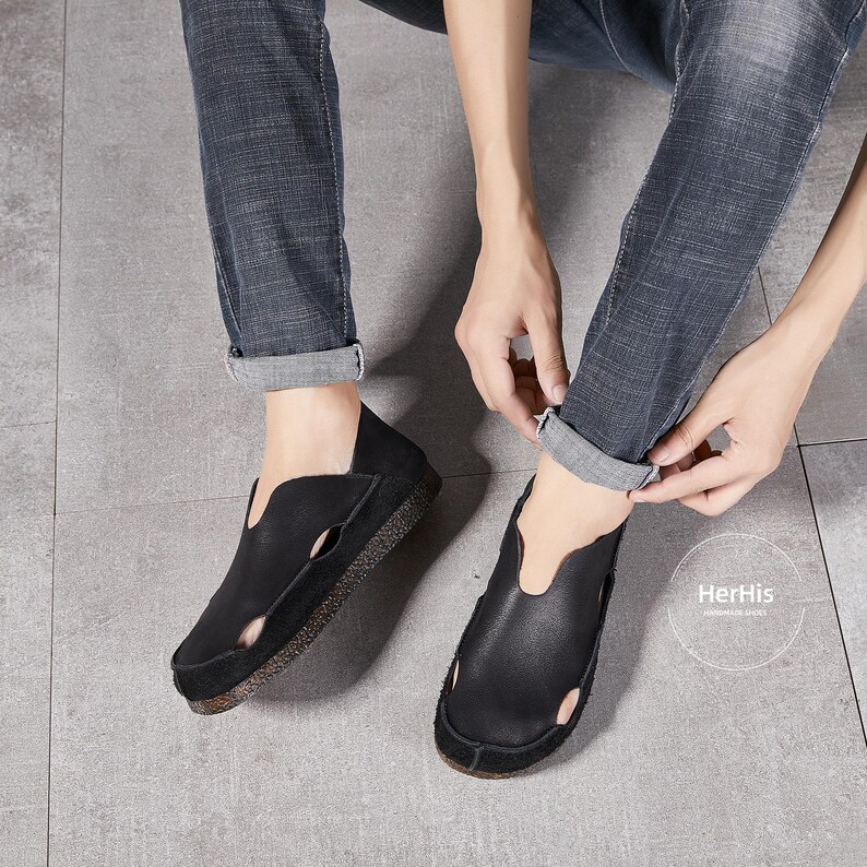 May include: A pair of black leather slip-on shoes with a cut-out design. The shoes have a rubber sole and are worn by a person wearing jeans.