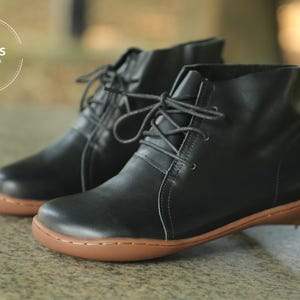 May include: A pair of black leather ankle boots with laces. The boots have a rounded toe and a brown rubber sole. HerHis Handmade Shoes is printed on the side of the boots.