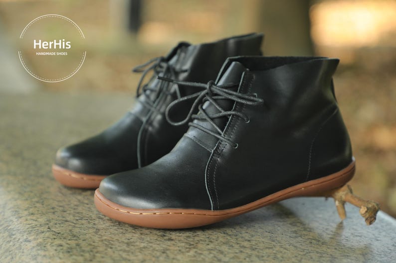 May include: A pair of black leather ankle boots with laces. The boots have a rounded toe and a brown rubber sole. HerHis Handmade Shoes is printed on the side of the boots.
