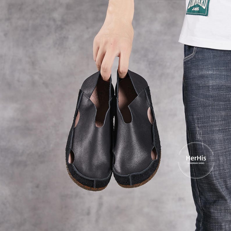 May include: A pair of black leather slip-on shoes with cutouts. The shoes have a suede sole and are held in a person's hand.