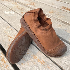 Op de afbeelding: Een paar bruine leren instappers met een textuurzool. De schoenen hebben een ronde neus en een gerimpelde kraag. De merknaam "HerHis Handmade Shoes" is zichtbaar op de achtergrond.