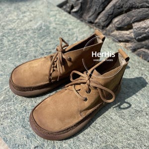 May include: A pair of brown leather ankle boots with laces. The boots have a rounded toe and a thick sole. The text "HerHis HANDMADE SHOES" is visible on the right boot.
