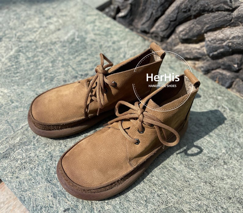 May include: A pair of brown leather ankle boots with laces. The boots have a rounded toe and a thick sole. The text "HerHis HANDMADE SHOES" is visible on the right boot.