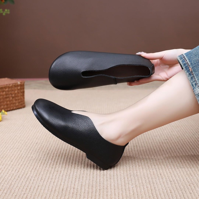 Op de afbeelding: Zwarte leren ballerina's met een ronde neus en een lage hak. E&eacute;n schoen wordt omhoog gehouden, wat het ontwerp laat zien. De andere schoen zit aan een voet. De schoenen hebben een getextureerd oppervlak.