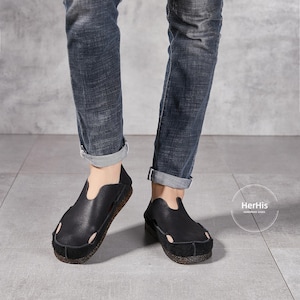 May include: A pair of black leather slip-on shoes with a simple design. The shoes have a pointed toe and a low heel. The shoes are being worn by a person wearing blue jeans.