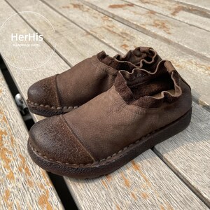 Op de afbeelding: Een paar bruin lederen instappers met een ronde neus en een gekruld kraagje. De schoenen hebben een getextureerd uiterlijk en een donkerbruine zool. De merknaam "HerHis Handmade Shoes" is zichtbaar op de achtergrond.