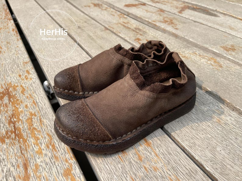 Op de afbeelding: Een paar bruin lederen instappers met een ronde neus en een gekruld kraagje. De schoenen hebben een getextureerd uiterlijk en een donkerbruine zool. De merknaam "HerHis Handmade Shoes" is zichtbaar op de achtergrond.
