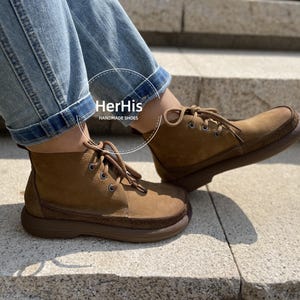 May include: A pair of brown leather ankle boots with laces. The boots have a thick sole and are made of suede. The boots are worn by a person wearing blue jeans. The text "HerHis HANDMADE SHOES" is visible on the boots.