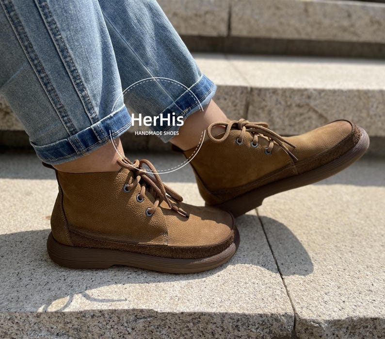 May include: A pair of brown leather ankle boots with laces. The boots have a thick sole and are made of suede. The boots are worn by a person wearing blue jeans. The text "HerHis HANDMADE SHOES" is visible on the boots.