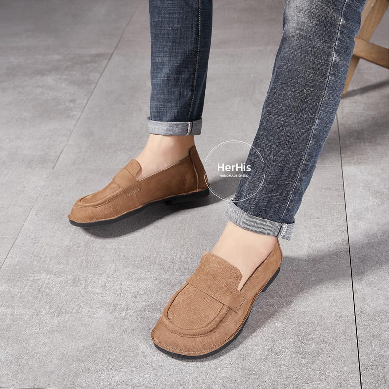 May include: A pair of brown leather loafers with a suede finish. The loafers have a simple design with a rounded toe and a low heel. The shoes are worn by a person wearing blue jeans.