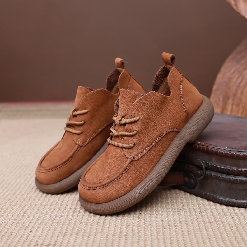 May include: A pair of brown leather ankle boots with a rounded toe and a lace-up closure. The boots have a thick, brown sole and a textured upper. The background is a neutral colour.