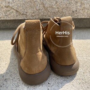 May include: A pair of brown leather ankle boots with a lace-up closure. The boots have a textured surface and a rounded toe. The brand name "HerHis HANDMADE SHOES" is printed on the side of the right boot.