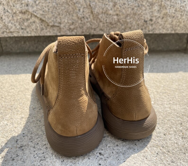 May include: A pair of brown leather ankle boots with a lace-up closure. The boots have a textured surface and a rounded toe. The brand name "HerHis HANDMADE SHOES" is printed on the side of the right boot.