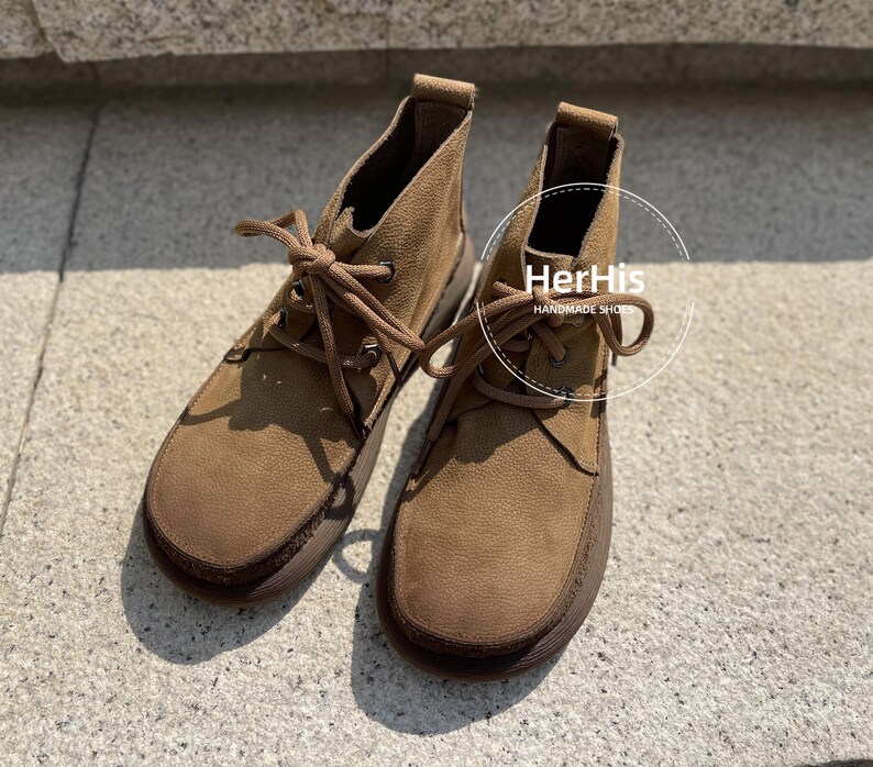 May include: A pair of brown suede ankle boots with laces. The boots have a rounded toe and a thick sole. The text "HerHis Handmade Shoes" is visible on the right boot.