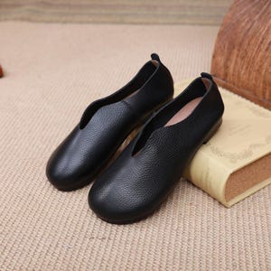 Op de afbeelding: Een paar zwarte leren ballerina's met een diepe V-vormige vamp. De schoenen hebben een getextureerd oppervlak en een ronde neus. Ze worden gepresenteerd op een beige oppervlak naast een boek.