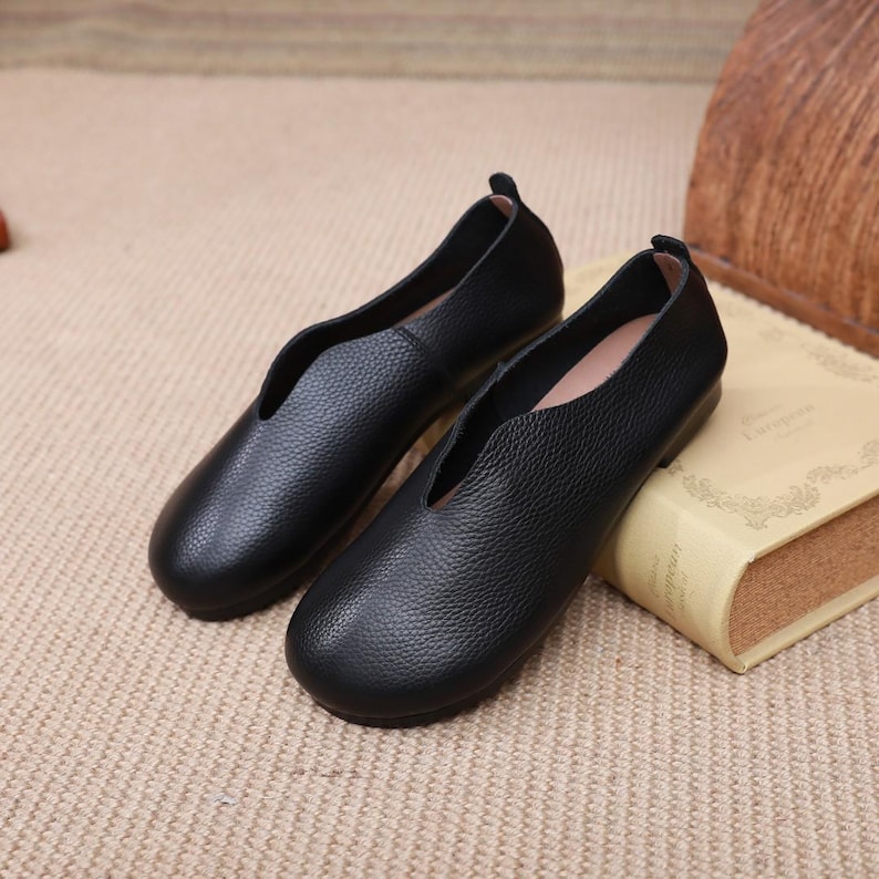 Op de afbeelding: Een paar zwarte leren ballerina's met een diepe V-vormige vamp. De schoenen hebben een getextureerd oppervlak en een ronde neus. Ze worden gepresenteerd op een beige oppervlak naast een boek.