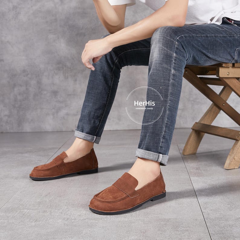 May include: A pair of brown suede loafers with a simple design. The loafers have a black rubber sole and a leather upper. The shoes are worn by a person sitting on a wooden stool. The text "HerHis Handmade Shoes" is visible on the image.