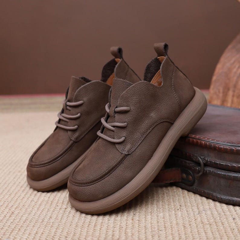 May include: A pair of brown leather shoes with laces. The shoes have a rounded toe and a thick, light brown sole. The shoes are made of a soft, textured leather and have a pull tab at the back.