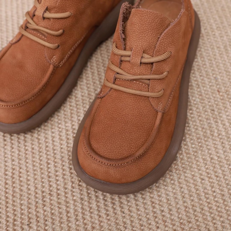 May include: Close-up of two brown leather shoes with matching laces. The shoes have a rounded toe and a thick, brown sole. The leather has a textured appearance. The shoes are on a neutral-coloured, textured surface.