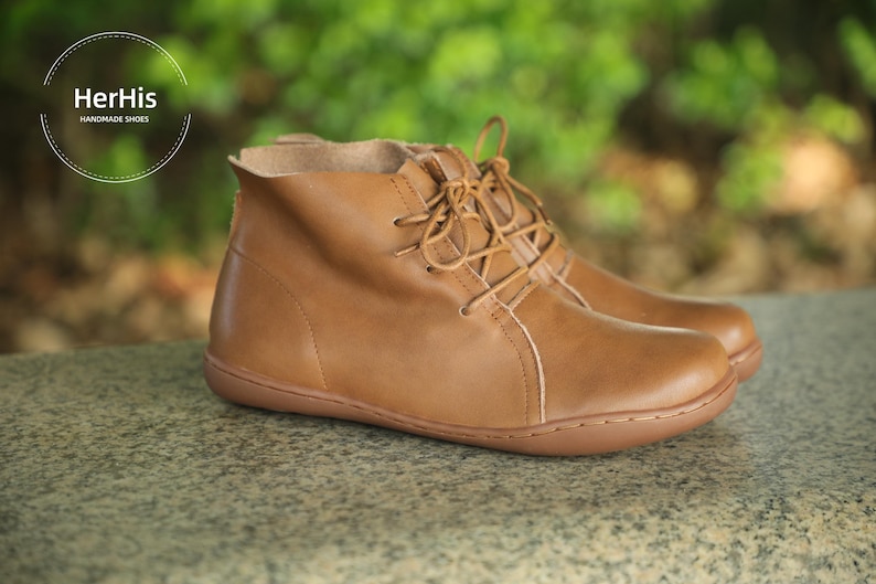 May include: Brown leather ankle boots with laces. The boots have a rounded toe and a slightly raised heel. The text "HerHis HANDMADE SHOES" is visible at the top of the image.