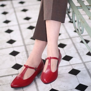 May include: A pair of red leather Mary Jane flats with a T-strap design. The shoes have a pointed toe and a small buckle closure. The image shows the shoes on a black and white tiled floor.