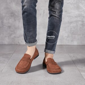 May include: Brown suede leather loafers with a slip-on design. The loafers have a rounded toe and a slightly raised heel. The shoes are worn by a person wearing blue jeans. The text "HerHis HANDMADE SHOES" is visible on the shoes.