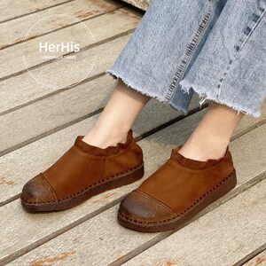 Op de afbeelding: Bruine leren schoenen met een ronde neus en een textuurzool. De schoenen hebben een gerimpelde kraag en zijn aan de randen gestikt. De merknaam "HerHis HANDMADE SHOES" is zichtbaar.