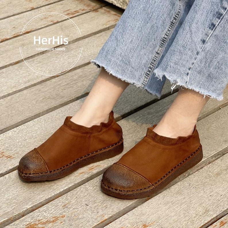 Op de afbeelding: Bruine leren schoenen met een ronde neus en een textuurzool. De schoenen hebben een gerimpelde kraag en zijn aan de randen gestikt. De merknaam "HerHis HANDMADE SHOES" is zichtbaar.