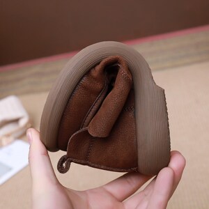 May include: Close-up of a brown leather shoe, demonstrating its flexibility. The shoe has a textured leather upper and a thick, light brown sole. The shoe is bent to show its pliable nature.