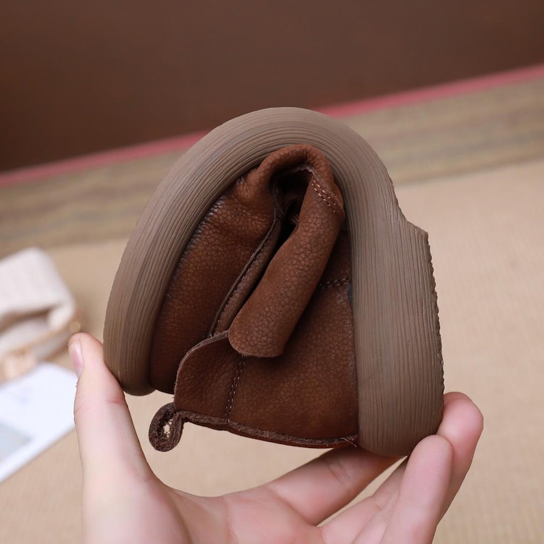 May include: Close-up of a brown leather shoe, demonstrating its flexibility. The shoe has a textured leather upper and a thick, light brown sole. The shoe is bent to show its pliable nature.