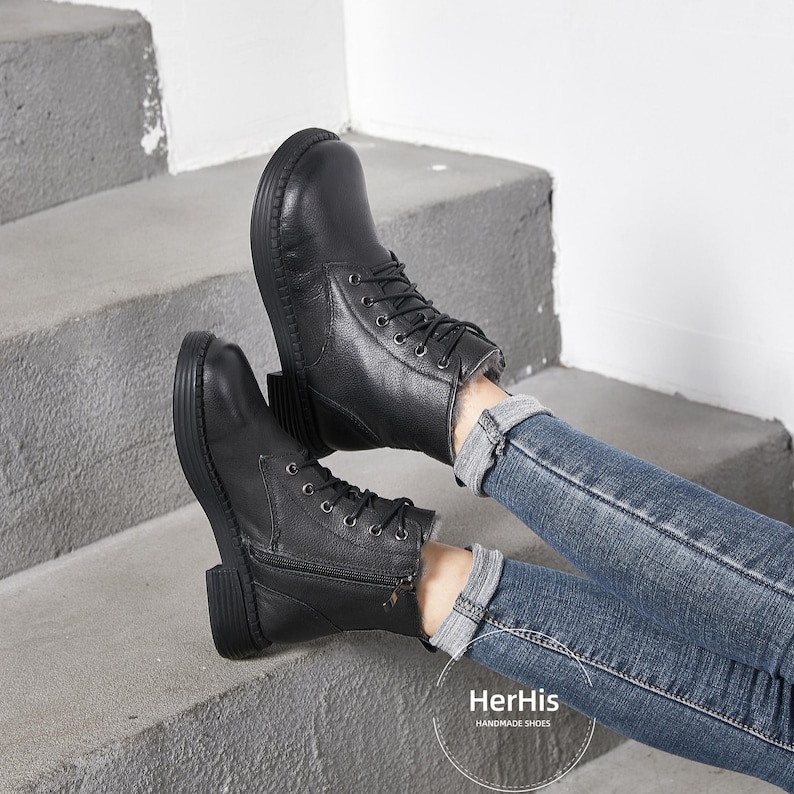 May include: A pair of black leather ankle boots with laces and zippers. The boots are worn by a person wearing blue jeans. The text "HerHis HANDMADE SHOES" is visible on the bottom of the image.