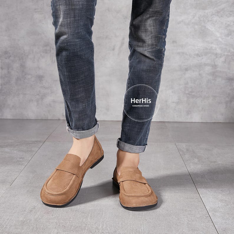 May include: A pair of brown leather loafers with a simple design. The loafers are worn by a person wearing blue jeans.