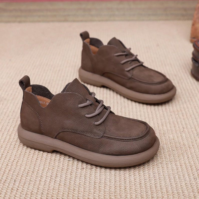 May include: A pair of brown leather shoes with a rounded toe and a thick sole. The shoes have laces and a low-cut design. The shoes are made of a textured leather material and are a neutral brown colour.